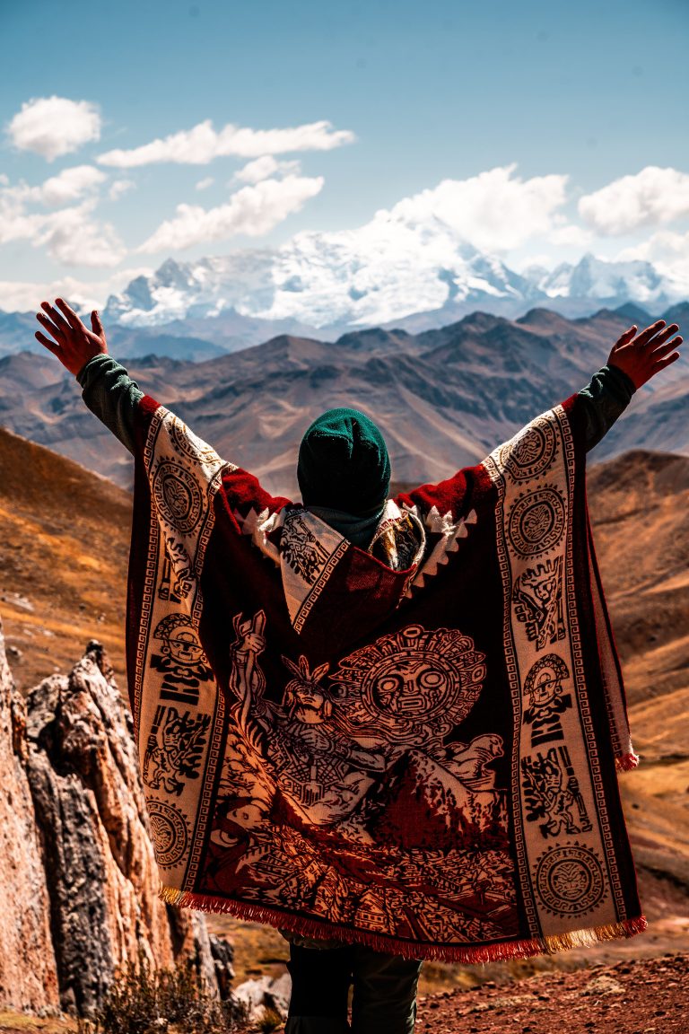 A person in traditional clothing with arms raised, standing on a mountain overlooking a snowy peak and a clear sky.